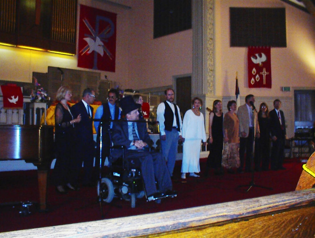 12 people singing on a stage. Kevin is in front of the rest, in a top hat, at a microphone. Two to the right in a long white dress is Nora Fox (white woman; short blond hair).