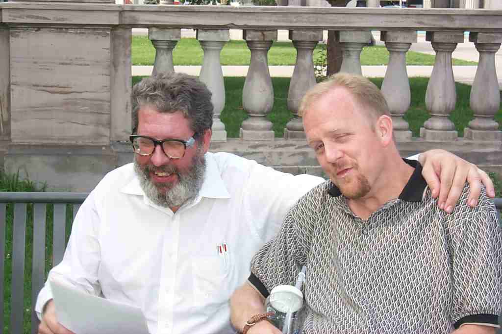 Dan Wilson (white man; salt & pepper hair and beard; white shirt) with his arm around Kevin (polo-style t-shirt).