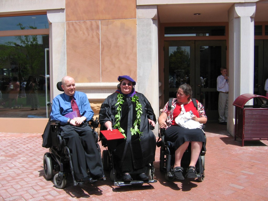 Kevin (blue shirt; red tie); Carrie Ann Lucas (white-presenting woman; wheelchair; wearing graduation cap and gown holding diploma in a red folder); Julie Reiskin (white woman; wheelchair; red shirt, black skirt).