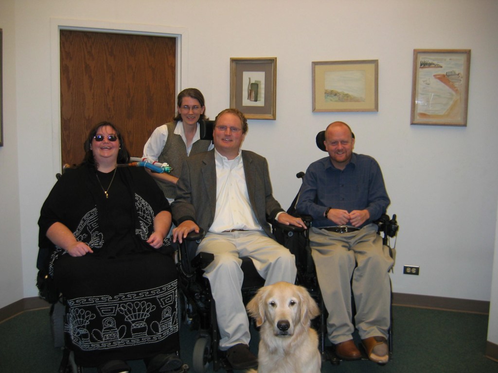 Four people and a dog posing in an office. Carrie Ann Lucas (white-presenting, large woman in wheelchair, brown hair, sunglasses, grinning); Amy Robertson (white woman; no wheelchair; brown hair; glasses); Tim Fox (white man; blond hair; wheelchair; sports coat); Kevin (wheelchair; blue shirt); and Chinook the golden retriever in the foreground.