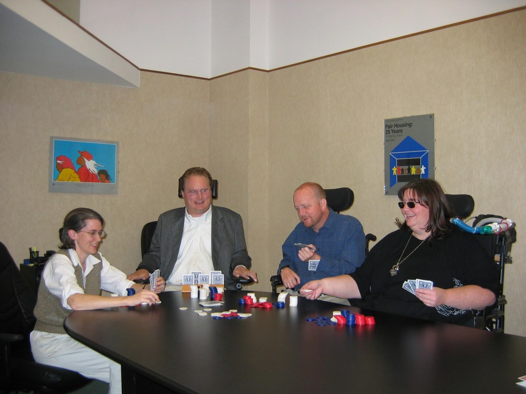 Four people posing playing poker an office table. Amy Robertson (white woman; no wheelchair; brown hair; glasses); Tim Fox (white man; blond hair; wheelchair; sports coat); Kevin (wheelchair; blue shirt); Carrie Ann Lucas (white-presenting, large woman in wheelchair, brown hair, sunglasses, grinning). On the table in front of us are poker chips and cards.