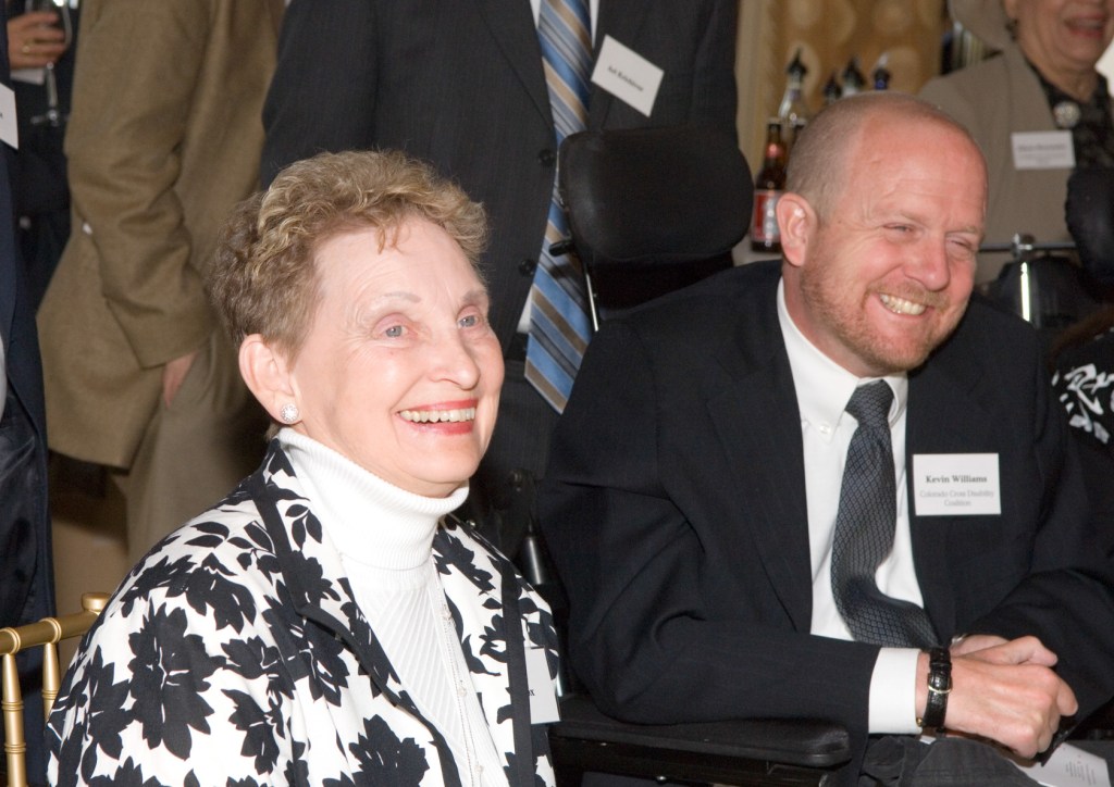 Kevin in a suit with a name tag, grinning/laughing. To his right, Nora Fox (white woman, short curly blond hair, black and white flowered jacket) also smiling.