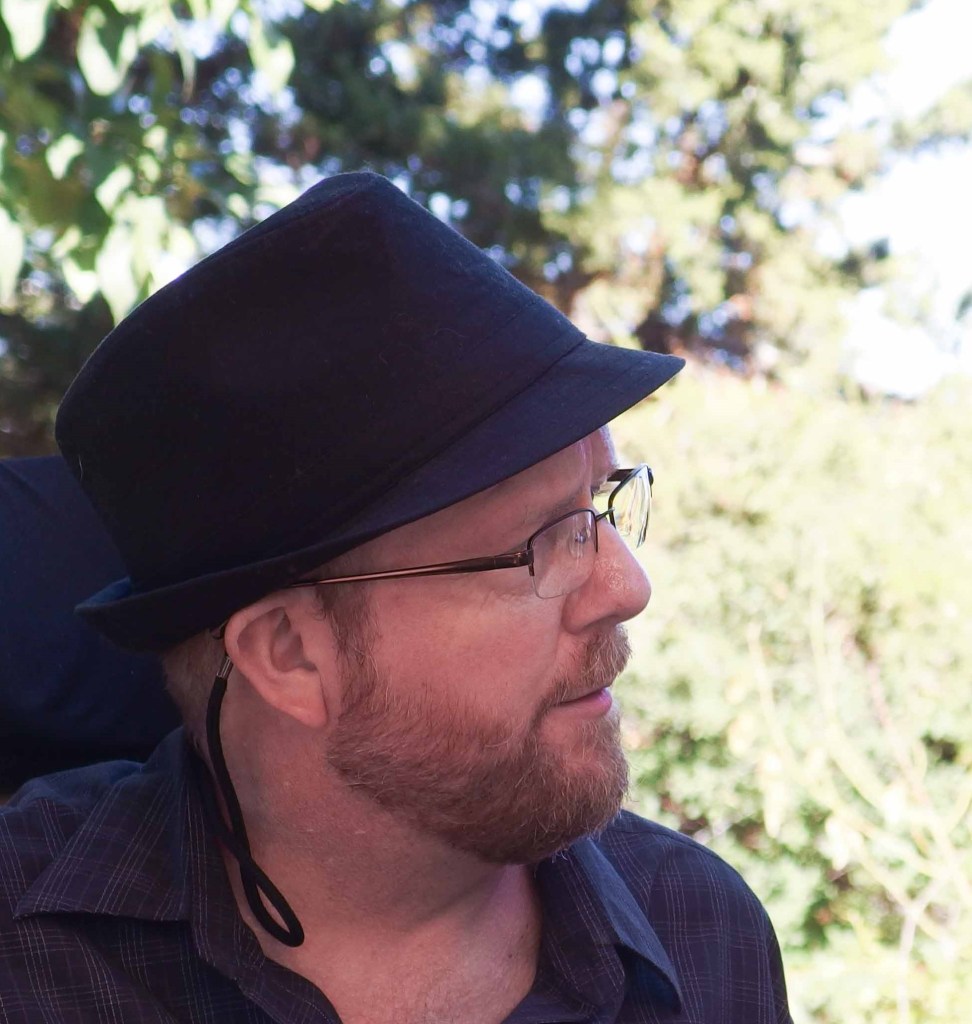 Kevin (white man; glasses; jaunty hat, short red beard) looking to the right of the frame, with a bemused expression, and, as always, preparing to talk.