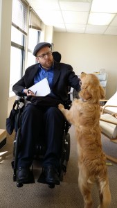 Kevin (white man; short red beard; glasses; flat cap; using wheelchair) playing with a golden retriever who has her paws up on his knees.