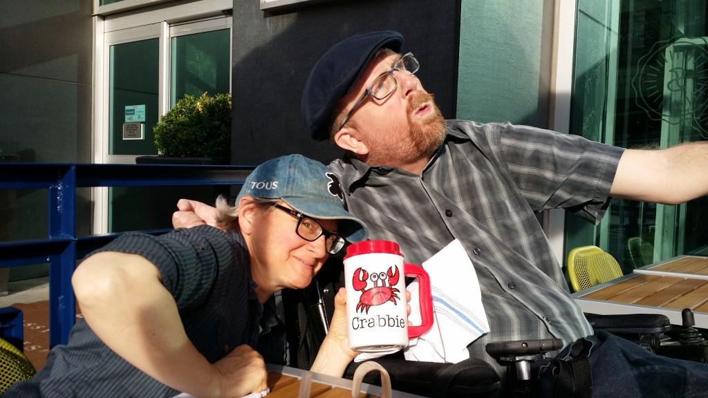 Kevin in a flat cap gesturing wit his left hand and appearing to sing; to his right leaning into him is Cara Reimann (white woman, denim ballcap, glasses) holding a mug that reads "Crabbie" with a cartoon crab.