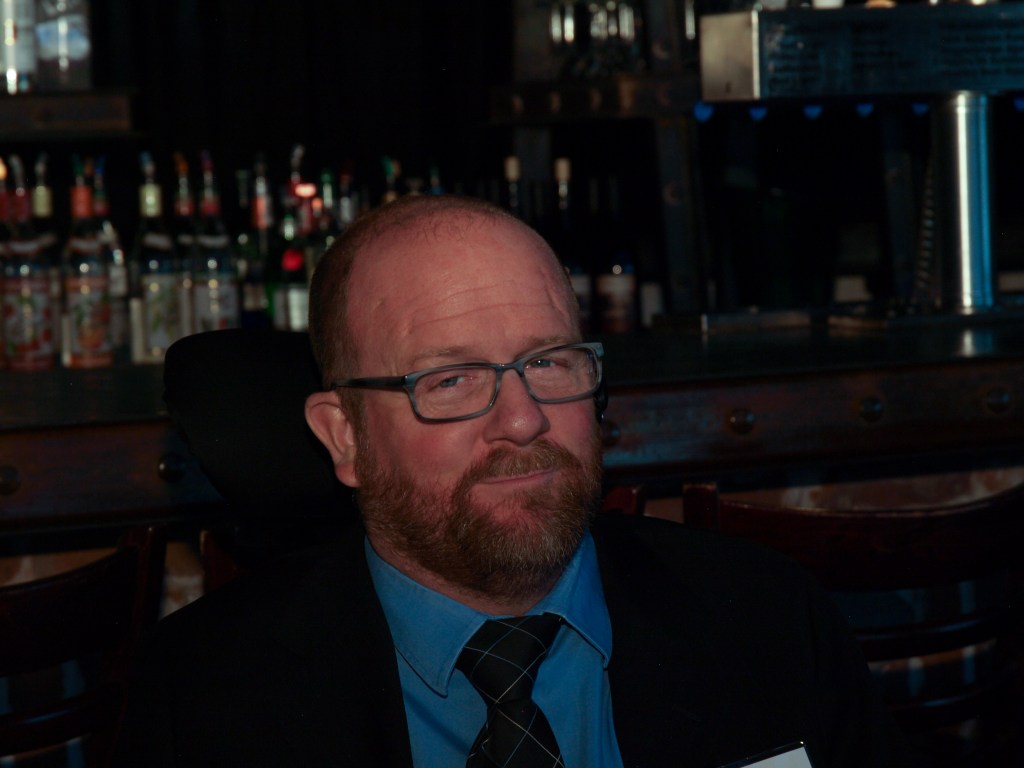 Kevin at a CCDC event, trying to look serious with a bar full of liquor bottles in the background.