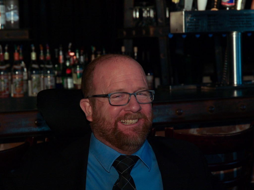 Kevin at a CCDC event, grinning with a bar full of liquor bottles in the background.