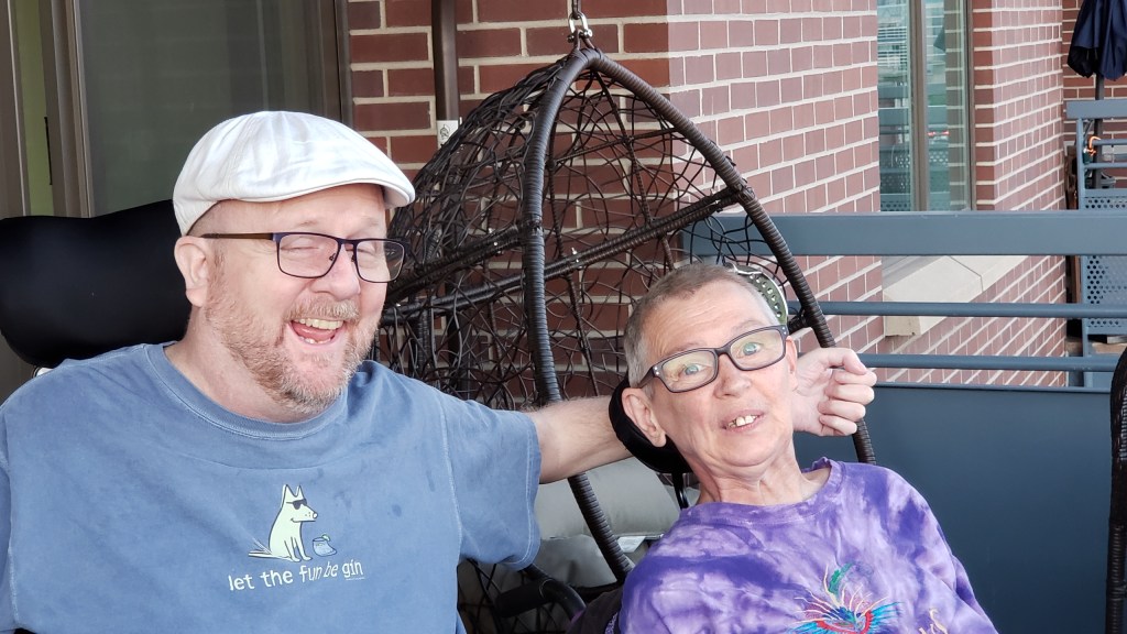Kevin (flat cap; t-shirt with cartoon dog reading "let the fun begin"); has his arm around Robin Stephens (white woman; glasses; very short hair; tie-dyed t-shirt; wheelchair) on the balcony of an apartment building.